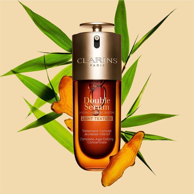Een flacon Clarins Double Serum Light Texture van 70 ml tussen kurkuma en Proven&ccedil;aals pijlriet tegen een oranje achtergrond.